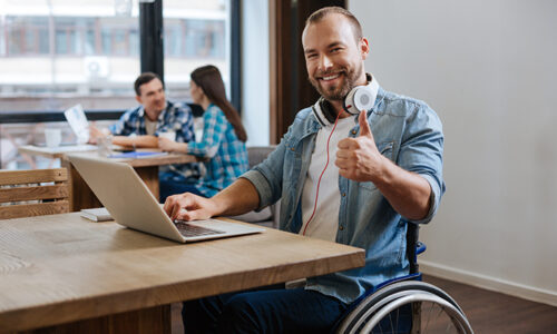 Un homme en fauteuil roulant souriant faisant un pouce levé tout en travaillant sur un ordinateur